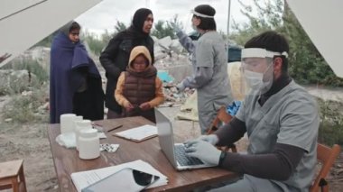 Erkek arkadaşı dizüstü bilgisayara not alırken, mülteci erkek, kadın ve çocukların sıcaklığını ölçen koruyucu elbiseli kadın sağlık görevlisinin orta boy fotoğrafı.