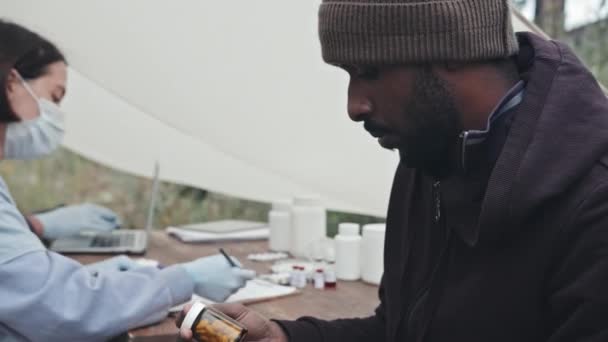 テントシティで医療援助を行う医療従事者のチームによって与えられた丸薬の瓶を保持しているアフリカ系アメリカ人難民の男性の中程度の閉鎖 — ストック動画