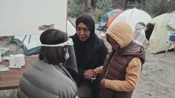 難民キャンプで小さな娘を調べながら 黒いヒジャーブのイスラム教徒の女性の話を聞く顔の盾の女性医療従事者の中程度の閉鎖 — ストック動画