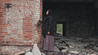 Yıkılmış evin harabelerinde kameraya bakarak duran tesettürlü Müslüman kadının tam portresi.