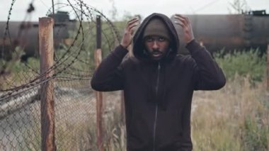 Afro-Amerikalı mülteci bir adamın orta boy portresi. Kapüşon takıyor, dışarıda dikenli tellerin yanında duruyor.