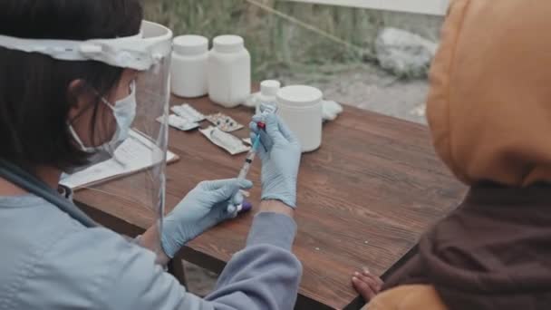 顔のシールドの女性医療従事者の後見人のメディア閉鎖は テントの街で小さな難民の女の子をワクチンで準備中に注射器を取得 — ストック動画