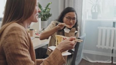 Orta yavaş çekimde genç Asyalı kadın ve ofis arkadaşları öğle arasında erişte yiyip iş yerinde sohbet ediyorlar.