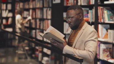 Genç Afro-Amerikan erkek profesörün modern kütüphane kitaplarında durup kameraya bakarken çekilmiş belden yukarı, yavaş çekimleri.