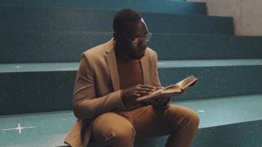 Orta yavaş çekimde Afro-Amerikan erkek üniversite öğrencisi akıllı günlük giysiler ve gözlük takıyor. Kapalı alandaki merdivenlerde oturmuş kağıt kitap okuyor.