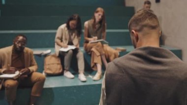 Modern erkek profesörün arka plan yakın plan çekimleri, koridordaki turkuaz merdivenlerde otururken çeşitli genç ve yetişkin öğrencilerin cevap vermek için el kaldırmalarına konferans veriyor.