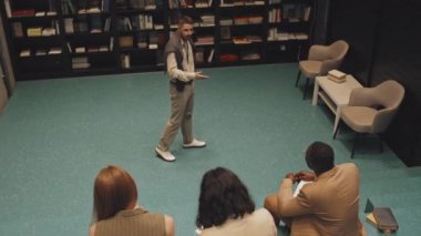 Modern kütüphanede merdivenlerde oturan çok ırklı öğrencilere akıllı günlük kıyafet giymiş çağdaş erkek üniversite profesörünün yavaş çekimini yukarıdan takip ederek ders veriyor.