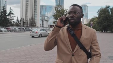 Beyaz giysili ve omuz çantalı özgüvenli Afro-Amerikan iş adamının şehir merkezinde yürürken telefon görüşmesi yaptığı belden yukarı ağır çekim.