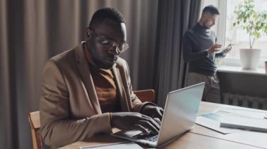 Ofiste oturan iş belgelerini yönetirken dizüstü bilgisayarında yazan ciddi bir Afro-Amerikan iş adamının belden aşağısı. İş ortağı arka planda dijital tablet kullanıyor