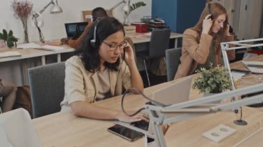 Kulaklık takan genç çağrı merkezi yöneticilerinin çalışma masalarında bilgisayar başında çalışırken orta yavaş çekimleri.