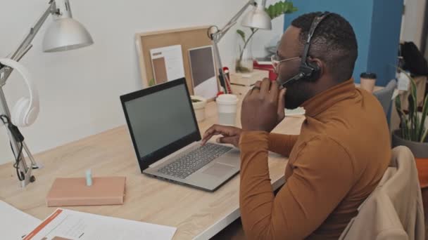 ヘッドセットでアフリカ系アメリカ人男性コールセンターの演算子のサイドビュー中低速ショットクライアントに話をし 現代のオフィスに座ってラップトップ上で入力 — ストック動画
