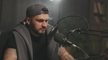 Kapaklı sakallı bir gencin podcast ya da radyo programında mikrofona konuşma görüntüsü.