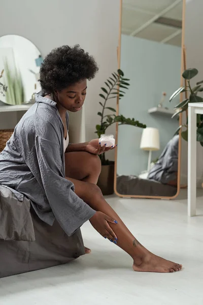 Mujer africana en ropa de casa gris aplicando crema en las piernas — Foto de Stock