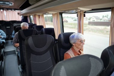 Beyaz saçlı, yaşlı, beyaz bir kadın otobüsün içinde, pencerenin kenarında oturuyor.