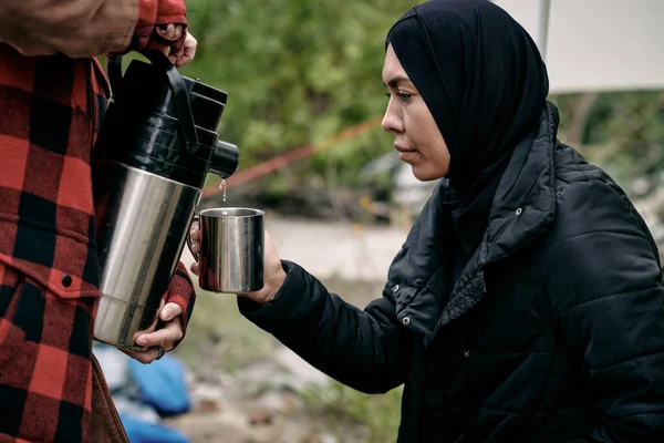 Joven musulmana vestida de negro sosteniendo taza mientras voluntaria vierte agua caliente —  Fotos de Stock