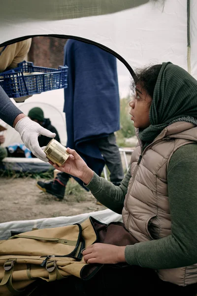 Голодная девушка-беженка берет консервы из перчаточной руки добровольца — стоковое фото