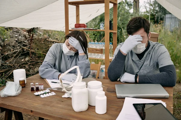 Twee jonge uitgeputte therapeuten in beschermende werkkleding aan tafel — Stockfoto