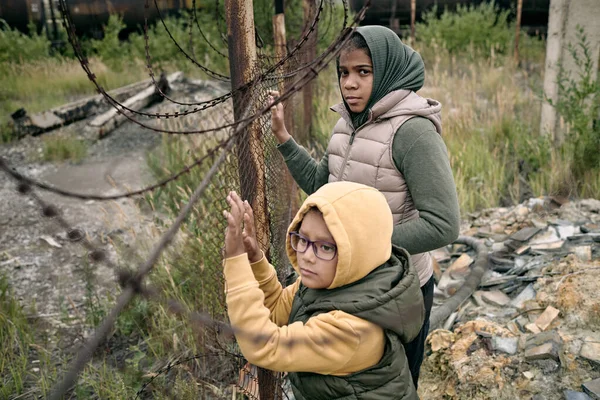 Dos niñas en sudaderas con capucha y chaquetas junto a la red que divide el campo de refugiados —  Fotos de Stock