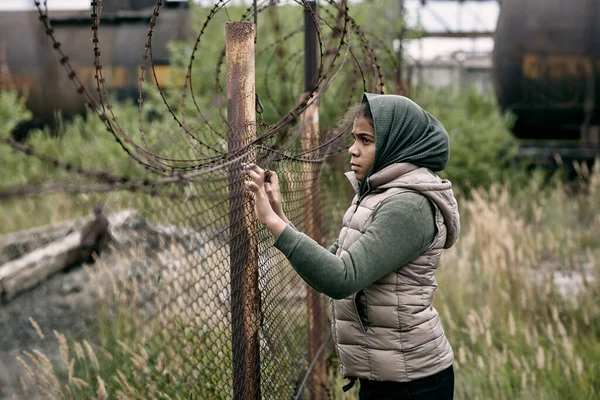Безпритульна дівчина в повсякденному одязі стоїть біля бар'єрного дроту паркану — стокове фото
