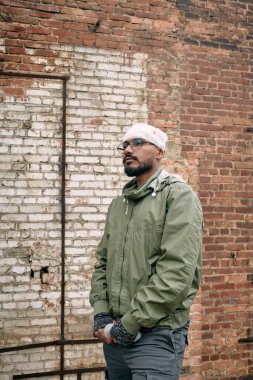 Kafasında bandaj olan genç adam tuğla duvarın yanında duruyor.