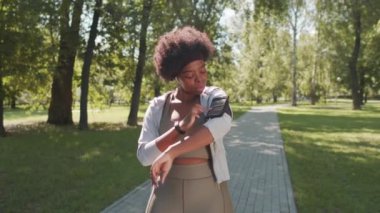 Genç Afro-Amerikan kadın spor kıyafetleri içinde, yaz günü parkta açık hava sporları antrenmanı yaparken kolluklarına bağlı akıllı telefon kullanırken orta yavaş bir çekim.