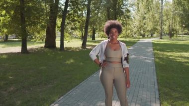 Spor giyim ve kol bandı takmış gülümseyen Afro-Amerikan kadının orta yavaş portresi. Spor antrenmanı sırasında elleri bağlı kameraya poz veriyor.