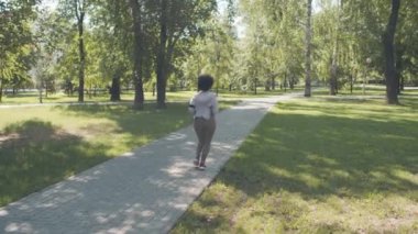 Sıkı spor kıyafetleri içinde, yaz günü parkta tek başına koşan Afrikalı Amerikalı bir kadının yavaş çekim yaptığı uzun bir dikiz görüntüsü.