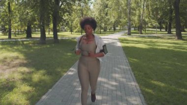Sıkı spor kıyafetleri içinde genç ve iri yarı Afro-Amerikan bir kadının güneşli bir günde parkta koşusunu bitirirken akıllı saatine bakıp gülümsemesinin yavaş çekimleri.