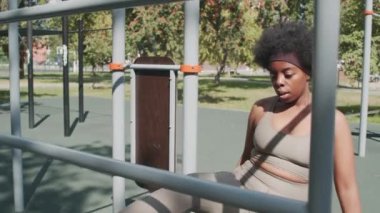 Orta yavaş çekimde, dar spor giysili, etkin Afro-Amerikan kadın güneşli bir günde tek başına ekipman eğitimi alarak esneme egzersizleri yapıyor.