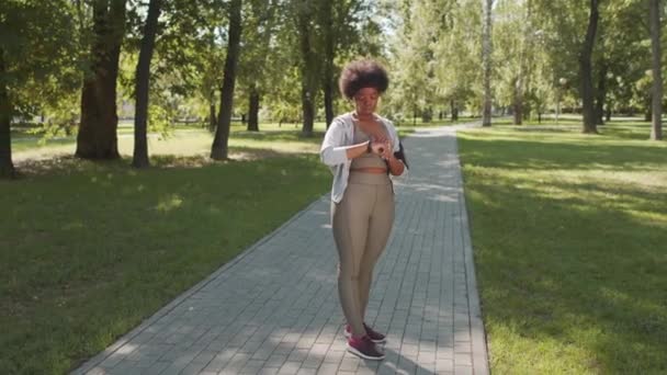 在阳光灿烂的日子里 身穿运动服的年轻非洲裔美国女子在公园慢跑时 在她聪明的身体上装上定时器 拍摄了全长的慢镜头 — 图库视频影像
