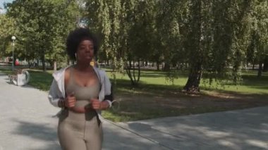 Güneşli bir günde parkta tek başına koşan, sıkı spor giysili, aktif, Afro-Amerikan bir kadının orta yavaş çekimini izliyorum.
