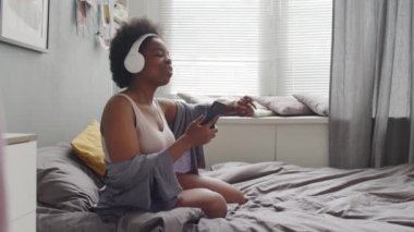 Orta yavaş çekimde, kulaklıkla müzik dinlerken sıradan iç çamaşırlı, dans eden ve şarkı söyleyen Afro-Amerikan genç mutlu kadın.