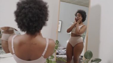 Yatak odasında aynanın önünde dikiz aynasının önünde duran, iç çamaşırlı, kıvrımlı, gülümseyen Afro-Amerikan kadının orta derece yavaş çekimi. Kendini sevme ve vücut pozitifliği kavramı