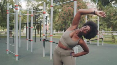 Orta yavaş çekimde, arka planda spor malzemelerine karşı esneme hareketleri yapan Afrikalı-Amerikalı genç bir kadın görülüyor.