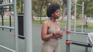 Spor salonunda spor salonunda spor yaparken dar spor giyim kuşamında içme suyu içen genç ve iri yarı bir Afro-Amerikan kadının orta yavaş çekimi.