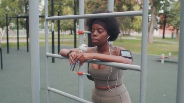 Spor giyimli ve kulaklıklı genç bir Afro-Amerikan kadının açık hava eğitimi sırasında spor tesisine yaslanırken, akıllı telefon için kol bandı takarken orta yavaş çekimleri.