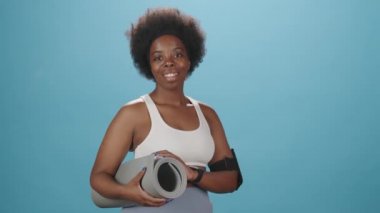 Dar spor giyim ve kolluk takmış gülümseyen Afro-Amerikan kadının orta boy bir portresi. Mavi arka planda yoga minderiyle kameraya poz veriyor.