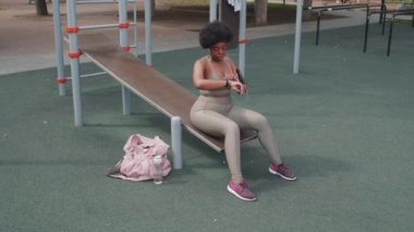 Spor salonundaki spor salonunda oturan ve spor sonrası dinlenen müzik dinleyen Afro-Amerikan genç ve iri yarı bir kadının tam boy bıçak yavaş çekimi.