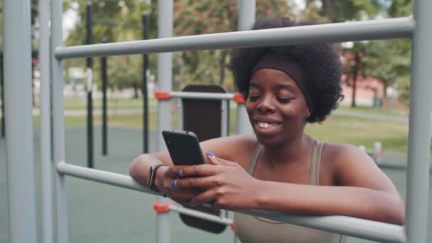 Střední zblízka s pomalým pohybem mladé usmívající se Afroameričanky v těsném sportovním oblečení pomocí smartphonu stojící na sportovním hřišti během outdoorového tréninku, opírající se o sportovní zařízení