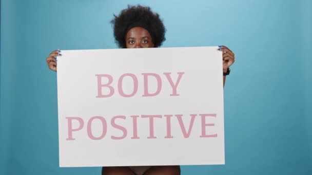 身着粉色内裤 手持印有身体阳性字样的大白海报 站在蓝色背景上的中号非洲裔美国女性照片 — 图库视频影像