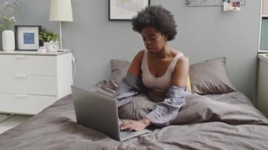 Rahat bir ev elbisesi içinde yumuşak bir yatakta oturup dizüstü bilgisayarında daktilo yazan Afro-Amerikan genç kadının orta yavaş portresi.