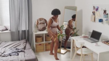 Yatak odasında aynanın önünde bant ölçüsüyle kalçalarını ölçen pembe iç çamaşırlı Afro-Amerikan bir kadının tam boy yavaş çekimi.