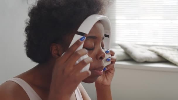 若いですアフリカ系アメリカ人女性の遅いクローズアップ上の白い無線ヘッドフォンを置きながら過ごす週末の朝一人でベッドで居心地の良い家 — ストック動画
