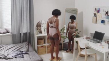 Dikiz aynasından, yatak odasında terazide duran ve vücudunu aynada inceleyen, iç çamaşırlı, Afro-Amerikan bir kadının tam boy yavaş çekimi.