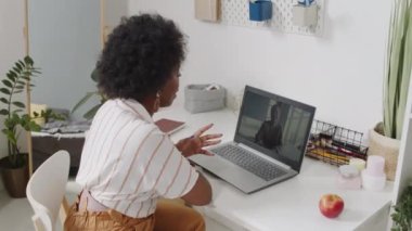 Genç ve çekici iş kadınının laptopta iş arkadaşlarıyla sohbet ederken çekilmiş, rahat bir dairede masada otururken çekilmiş dikiz görüntüsü.