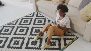 Modern apartmandaki koltukta sırtüstü uzanmış, elma yiyen ve dijital tabletleri karıştıran gündelik giysili genç Afro-Amerikan kadının yüksek açılı yavaş çekimi.