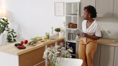 Orta yavaş çekimde, genç Afro-Amerikalı bir kadının dijital tabletten tarifine bakarken modern apartmanın mutfak masasında sağlıklı bir yemek hazırlarken...