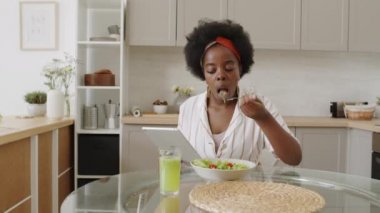 Orta yavaş çekimde, sağlıklı sebze salatası ve detoks içerken modern mutfakta yemek masasında dijital tablet kullanan Afro-Amerikalı genç bir kadın görülüyor.