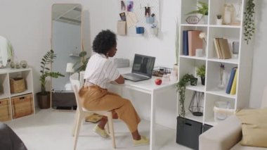 Genç, çekici, Afro-Amerikalı iş kadınının laptopta görüntülü konuşma yaparken, rahat bir dairede, masada otururken.
