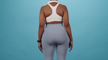 Kendine güvenen, spor giyimli Afro-Amerikan bir kadının mavi stüdyo arka planında kameraya poz verirken çekilmiş dikiz aynalı portresi.